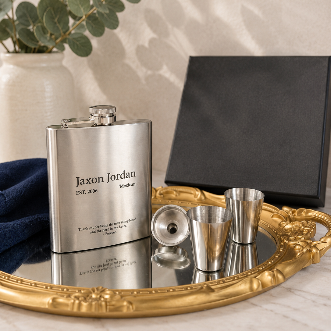 A personalized stainless steel flask with a matching metal decanter and two small metal tumblers arranged on an ornate gold tray, with a black presentation box and eucalyptus branches in the background.