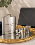A personalized stainless steel flask with a matching metal decanter and two small metal tumblers arranged on an ornate gold tray, with a black presentation box and eucalyptus branches in the background.